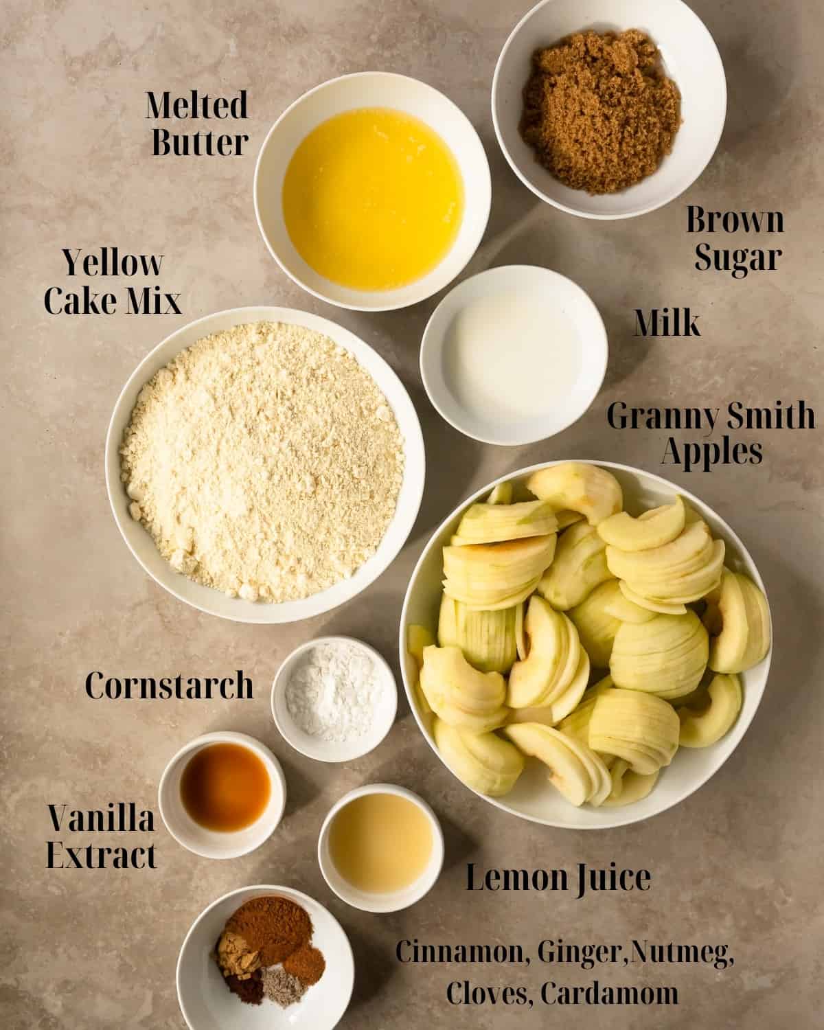 Apple Cake Mix Cobbler Ingredients in bowls. The main ingredients for this apple cobbler dump cake recipe are fresh baking apples such as granny smith, brown sugar, ground cinnamon, ginger, nutmeg, cloves and cardamom, lemon juice, cornstarch, yellow cake mix, butter and milk. 