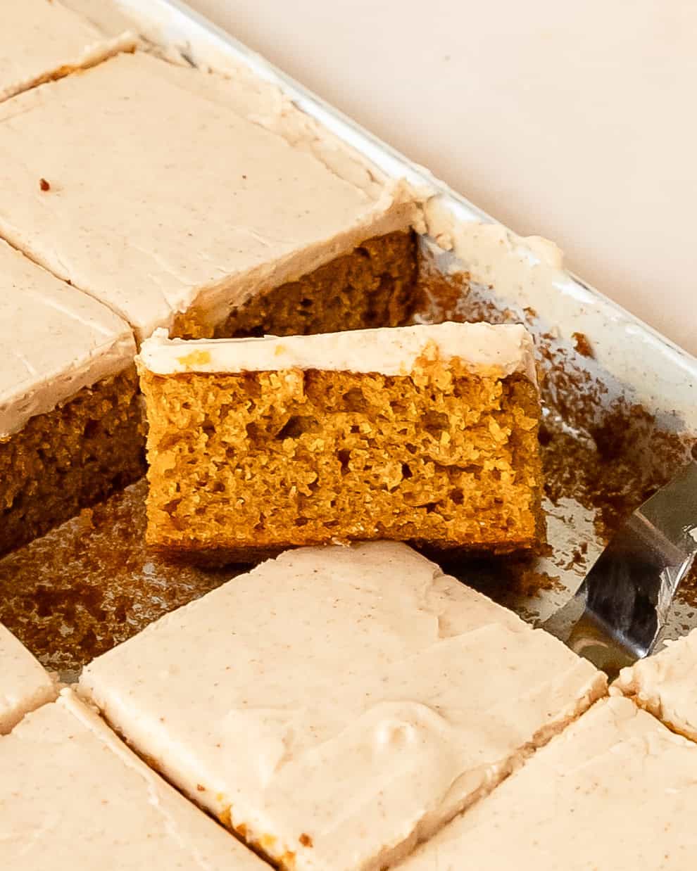 Slice of pumpkin cake bar on top of a pan of cream cheese frosted pumpkin bars.